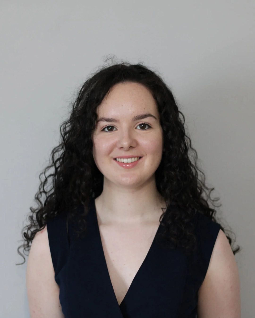 A young woman with long, curly dark hair is smiling at the camera. She is wearing a sleeveless dark top and is posed in front of a plain light gray background.